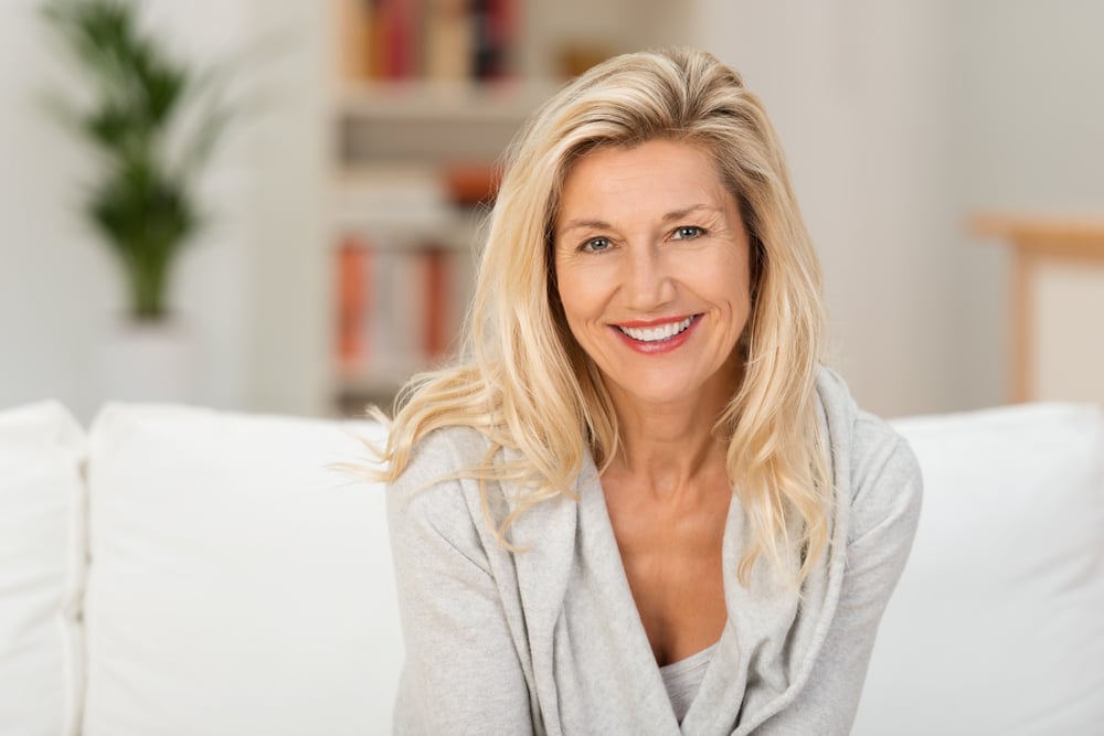 blond woman with a beaming smile sitting on a sofa at home looking at the camera